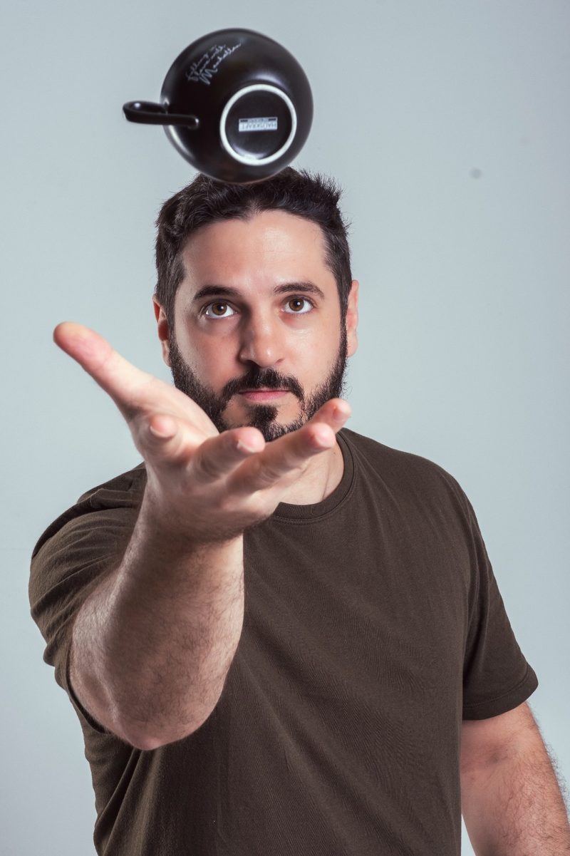 Sebastian Grau em retrato de estúdio, com uma xícara de café no ar acima da cabeça e a mão estendida para pegá-la